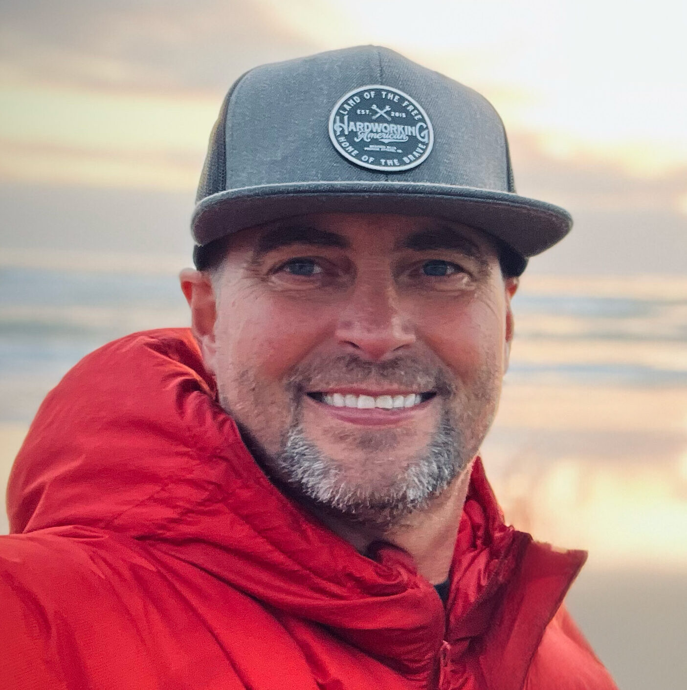 Smiling man in a red jacket and gray cap by the water at sunset.